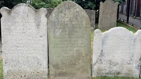 BBC / Steve Ladner Several historic gravestones stand in the former churchyard of St Mary's Church in Dover. At the centre is the memorial to Lieutenant James Hart, which records that he was killed at the Battle of Waterloo in 1815. The weathered stone is surrounded by other headstones, trees and railings.