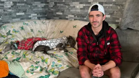 The picture shows a man named Shaun Reynolds pictured with two whippets. Shaun is sat to the right of the picture. He is wearing a white cap that is on his head backwards. He is also wearing a red and black tartan zip up jacket. His hands are resting on his knees and they are clasped together. He is smiling slightly. Behind him there is a grey brick wall. He is sat on a grey sofa. On top of the sofa is a duvet cover. It is beige with green, yellow and turquoise butterflies on it. Led on top of it is two whippets. They have grey fur. One has a red tartan dog coat the other is wearing a grey and white dog coat. The two dogs are sleeping.