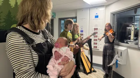 A baby, wearing a pink sleepsuit, sucking a pick dummy, sleeps in her mother's arms. They are listening to the musicians on the hospital ward who are playing a violin, harp and guitars.