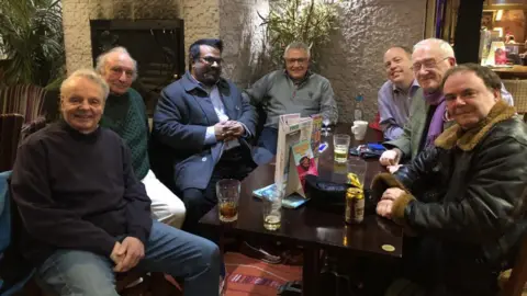 Vikki Irwin/BBC There are seven men dressed in casual clothing gathered around a table in a pub. 