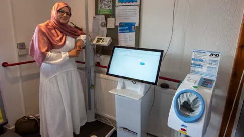Obaida is standing inside a surgery next to one of the new machines. She is wearing a hijab and a white dress and glasses. She is leaning on equipment next to the machine which is white with a computer screen above it.