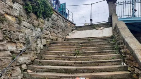 North Yorkshire Council The photograph shows a set of old stone steps leading upward between two stone walls. The stone walls on either side are weathered and aged, with visible moss and greenery growing in some crevices.