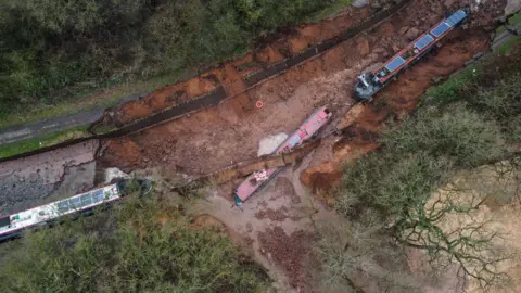 An aerial view of the breach of the Llangollen Canal at Whitchurch, showing a large brown-red scar in the ground where the bank and bed of the canal has washed away, with three boats left on the mud, one with a corrugated metal fence across it. Escaped water has flooded a neighbouring field.