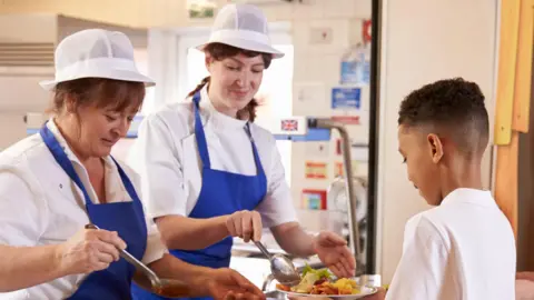 Two school cooks wearing white hats and blue aprons holding large spoons serving a plate full of food to a young pupil in a school canteen.