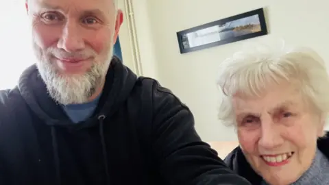 Tim has a grey beard and is wearing a black hoodie over a blue t-shirt. Next to him on the right is his mother Cymbria who has short grey hair and is wearing reddish-pink lipstick. She is also wearing a black coat over a grey knitted top and is smiling.