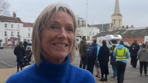 Jill Douglas wearing a blue turtleneck jumper. She has short blonde hair and is smiling. There is a group of people behind her standing in a car park.