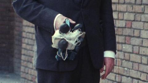 unidentifiable figure, head and shoulders out of shot, holds a Spider Criminal Apprehender in one, with the other hand by their side.  The contraption appears to have rubber stoppers and a sting bag within the primed device.