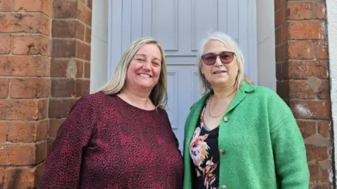 A woman with blonde hair and a dark red top beside a woman with grey hair, sunglasses and a green top in front of a white house door