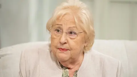 Sylvia Young, seen in 2015, wearing a cream cardigan and sitting on a sofa