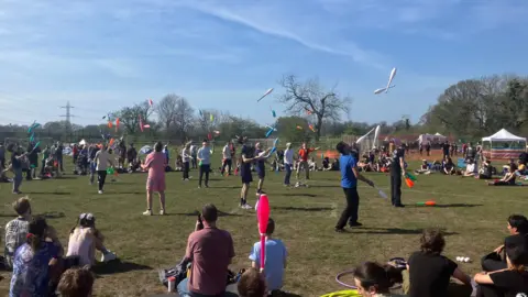 Dozens of jugglers throw clubs into the air. Surrounding them are hundreds of people sitting and watching them. They are sitting in a green field, with blue skies above.