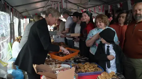 Dozens of people in a tent in front of a table filled with trays of food. On one side of the table are people serving and on the other are people looking to buy the food. One woman in a bandana looks and smiles at the camera (in the centre).
