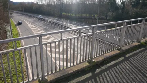 BBC The bridge has a metal railing fence either side of it as it spans the A442. The carriageways can be seen below with a vehicles on each side.