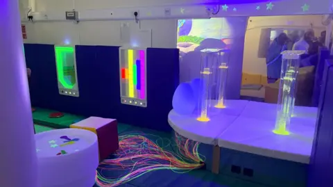 The Dawoodi Bohras UK A sensory room with purple lighting, padded floors, and illuminated features. There are tall bubbling light tubes, rainbow wall panels, and glowing fiber-optic strands create a colorful interactive space.
Soft seating, projected images, and cushioned surfaces make the room feel safe, soothing, and immersive.