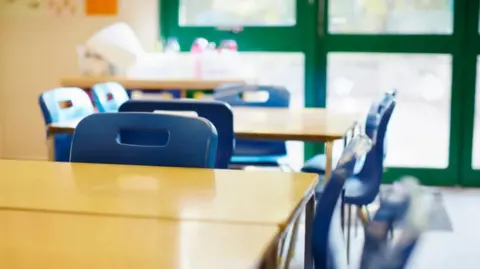 School desk in foreground in empty classroom with chair behind it and another desk and chair and window behind that