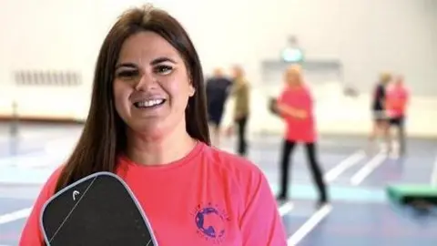 Christina is looking directly at the camera and is wearing the Suffolk Punch Pickleball T-shirt, which is bright pink with a blue logo. She is holding her pickleball paddle. There are other players in the background.