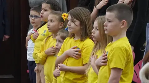 Children wearing yellow t-shirts stand in a horizontal line with adults behind them. They are singing adn hold a hand to their hearts.