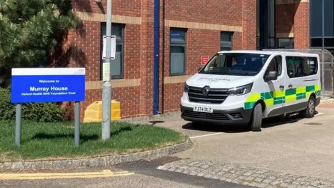 Outside a red brick building, a blue and white NHS sign reads "Welcome to Murray House".  An ambulance is parked alongside the building.