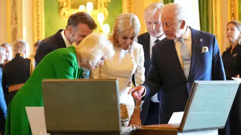 PA Media Queen Camilla, King Charles, French President Emmanuel Macron and his wife Brigitte view items laid out on a table that are obscured from view by two open cases which contain the items.