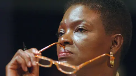 Close up of Baroness Valerie Amos with short hair taken from the side holding orange glasses with one of the arms touching her face above her lip. She looks serious. 