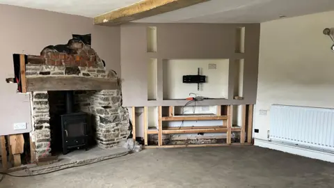 Photograph of what was Paul's living room. The far wall is beige and has patches of discolouration on it. The other two walls - one of which has been ripped out in the middle - are painted grey. The brick on the fireplace is exposed, and you can see cables trailing from where the TV has been taken off the wall. The floor is exposed, dusty concrete.