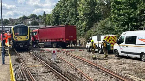 BBC/Julia Gregory A train with a lorry parked next to it on train tracks. Network Rail vans and people in hi-vis jackets are on the scene