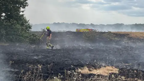 Laura Foster/BBC A single firefighter walking around a blackened field with wisps of smoke coming off it and a fire engine in the background.
