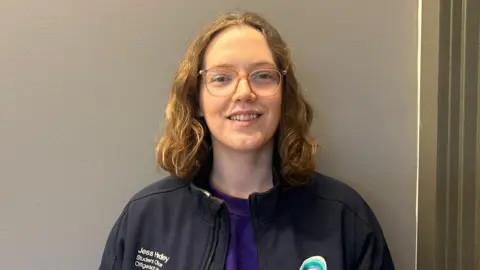 Jess smiles. She has brown glasses and brown curly hair. Her top is purple and she has on a black jacket with a Queen's Students' Union logo on it.