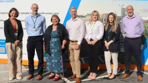 UHP Seven people, consisting of three men and four women, standing in front of the construction site smiling to camera