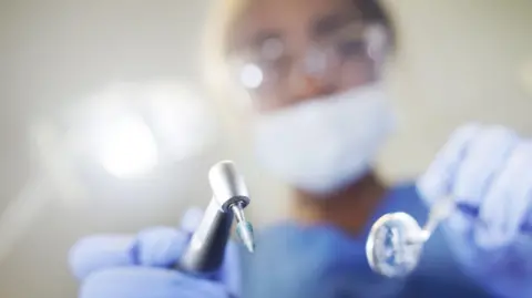 A dentist stands oversomeone with shiny metal tools. They are wearing glasses with a mask over their mouth. 