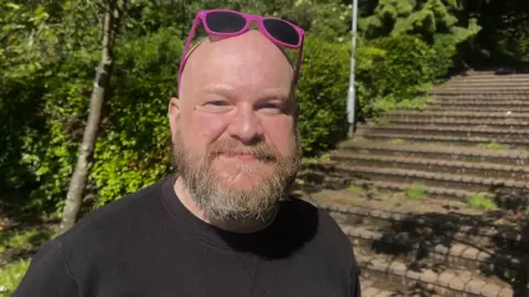 A man with a bald head, beard and pink sunglasses on top of his head in a black T shirt, standing in front of rows of terraced stone seats and some trees