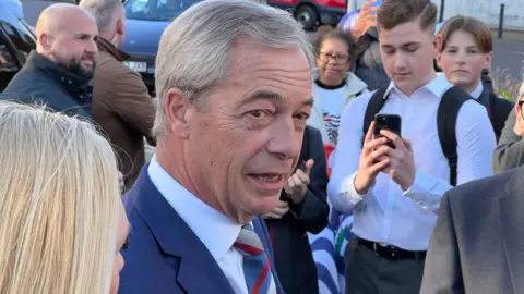 Nigel Farage close up. He is talking to people in a crowd. He is wearing a blue suit and shirt with a stripy tie. A young male wearing a white shirt with a rucksack is holding a smartphone in front of his chest - possibly taking a photo of Farage. There are security guards in the background.