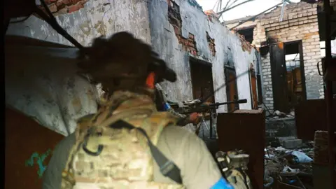 Conall Kearney Two unformed Ukrainian soldiers with guns stand outside a heavily bombed building