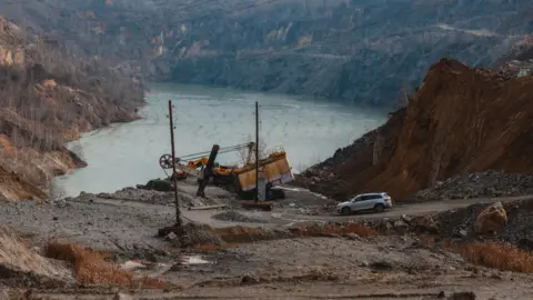 Mining machinery and a car at the open-pit Zavallya mine, with mountains and a lake in the background