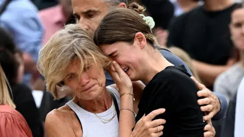 EPA Two woman are consoling one another as they attend a memoral at Bondi Beach in Sydney. The woman on the left is an older woman with short blonde hair. She is hugging a younger woman on her right, who is crying into her hand.