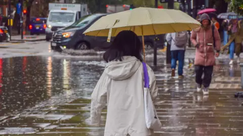 Getty Images The image shows a woman holding an umbrella in the rain with other people walking up and down the pavement. Cars and other vehicles can be seen driving through flooded streets in the background.