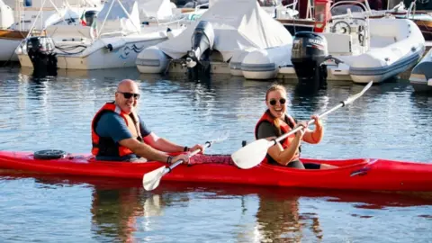 BBC/Studio Lambert a man and woman on a tandem kayak, holding their paddles out of the water and smiling at the camera