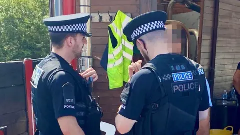 Two police officers in uniform are talking to a person whose face has been hidden from view. They are standing in front of a wooden building.