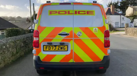 The back of a police van parked next to a stone wall in a residential area of Cornwall. The van had orange and yellow chevrons on it along with the word police written in large orange block capitals on a yellow background. 