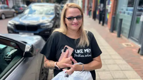 Becki Bowden/BBC A woman with blonde hair and sunglasses holds her mobile phone and shopping bags as she smiles into the camera. She is standing on a street with shops on the right and parked cars on the left.