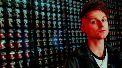 Look Mum No Computer stands in front of his 1,000 Oscillator Drone Synth, bathed in red light