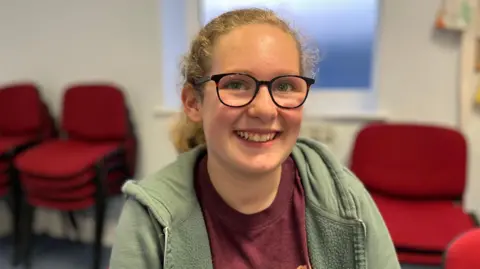A girl with blonde curly hair and glasses smiling at the camera. She is wearing a green hoodie and a burgundy t-shirt.