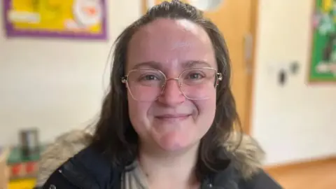 EMMA BAUGH/BBC Image shows Anna smiling and facing the camera. She is wearing glasses and a hooded jacket. In the background is the wall of a family hub with children's artwork on the wall. 