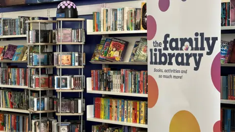 A tall banner reads "The Family Library: books, activities and so much more!" It is white with multicoloured spots and is standing in front of a bookshelf full of books.