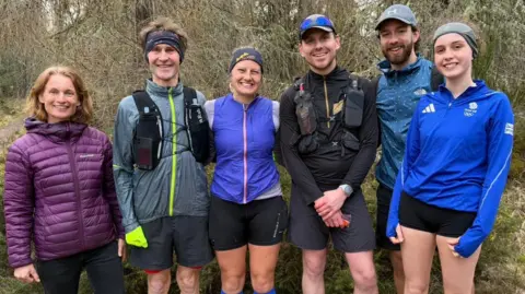 Freya Shepherd The photo taken in the woods, the trees are visible on the background. Freya is standing in the middle of the shot and her support runners are standing next to her. 
On her right hand side is a man in a grey running jacket and shorts, with black headband, and a woman with shoulder length fairly red hair, purple jacket and black leggings. 
On Freya's left hand side is a man in black top and shorts with a grey cap, next to him on his left stands another man in a blue jacket, grey cap, black shorts and his having a beard. And then, further left stands a woman in blue adidas quarter zip top, black shorts and grey headband. Everyone on the picture has big smile on their face.