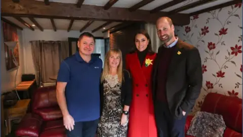 Princeandprincessofwales/Instagram (left to right) Butcher Kevin Kidner, Zsuzsanna, Catherine and William.
The four of them are stood in front of red sofas in a room with beams on the ceiling. Kevin is wearing a blue t-shirt and blue jeans, Zsuzsanna is wearing a leopard print dress under a black cardigan, Catherine is wearing a red coat with a daffodil pin on the left lapel, and William has blue trousers, a maroon jumper under a black blazer with a daffodil on the left lapel.