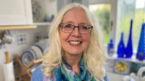 Lucy Gardner souriante dans la cuisine. Elle a une audience blonde jusqu'aux épaules et des lunettes.