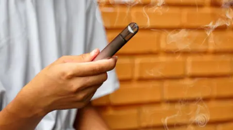 A young person holds a black and grey vape pen with smoke unfurling from the top. He is standing against a brick wall and his face is not visible, only a light grey t-shirt and his hand.