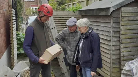A man and a woman are standing next to another man, looking t a box he is holding. The box is wooden and made for swifts, with a small hole in the corner. He is wearing a red hard hat and there is a ladder leaning onto the side of a house
