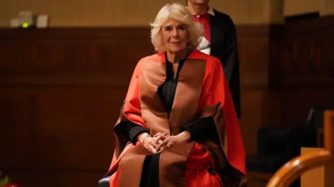 Queen Camilla's latest event saw her presented with an honorary doctorate on Wednesday. She is sitting on a chair wearing a clothes and a red and brown robe on top