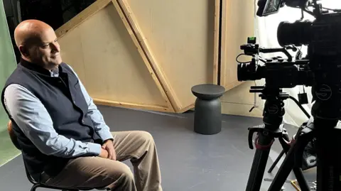 A side view of Sanjeen Payne-Kumar sat on a chair looking into the lens of a camera